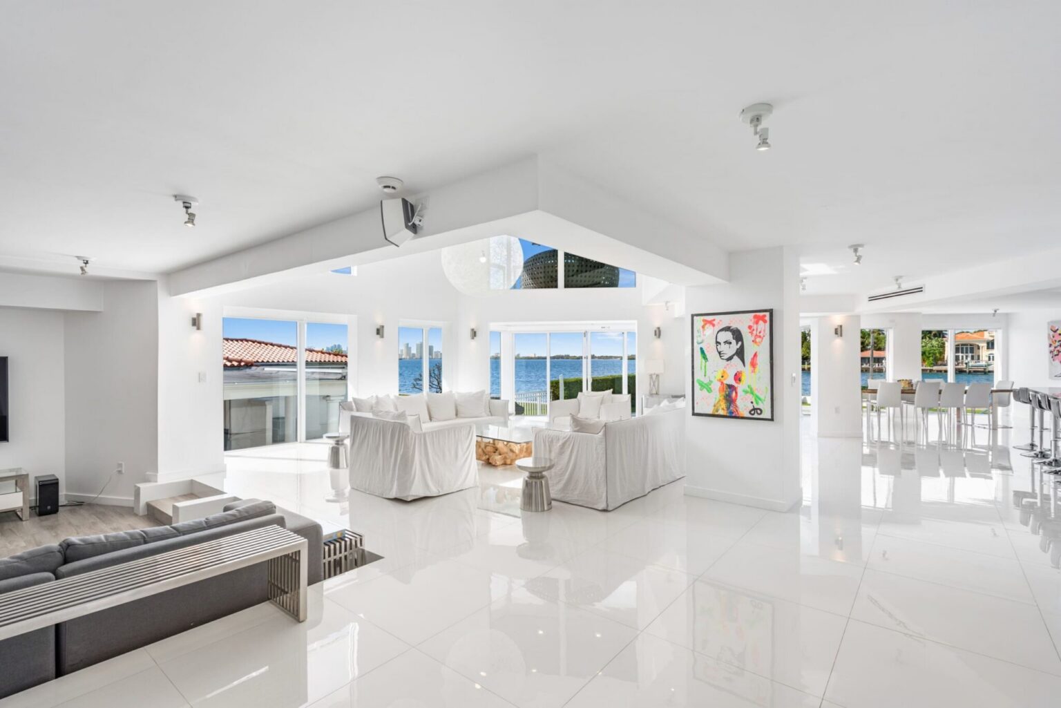 Expansive open-concept great room featuring multiple lounge seating areas, vaulted ceilings, glossy white tile flooring, contemporary artwork, large windows, and seamless connectivity between the media lounge, dining area, and waterfront living space