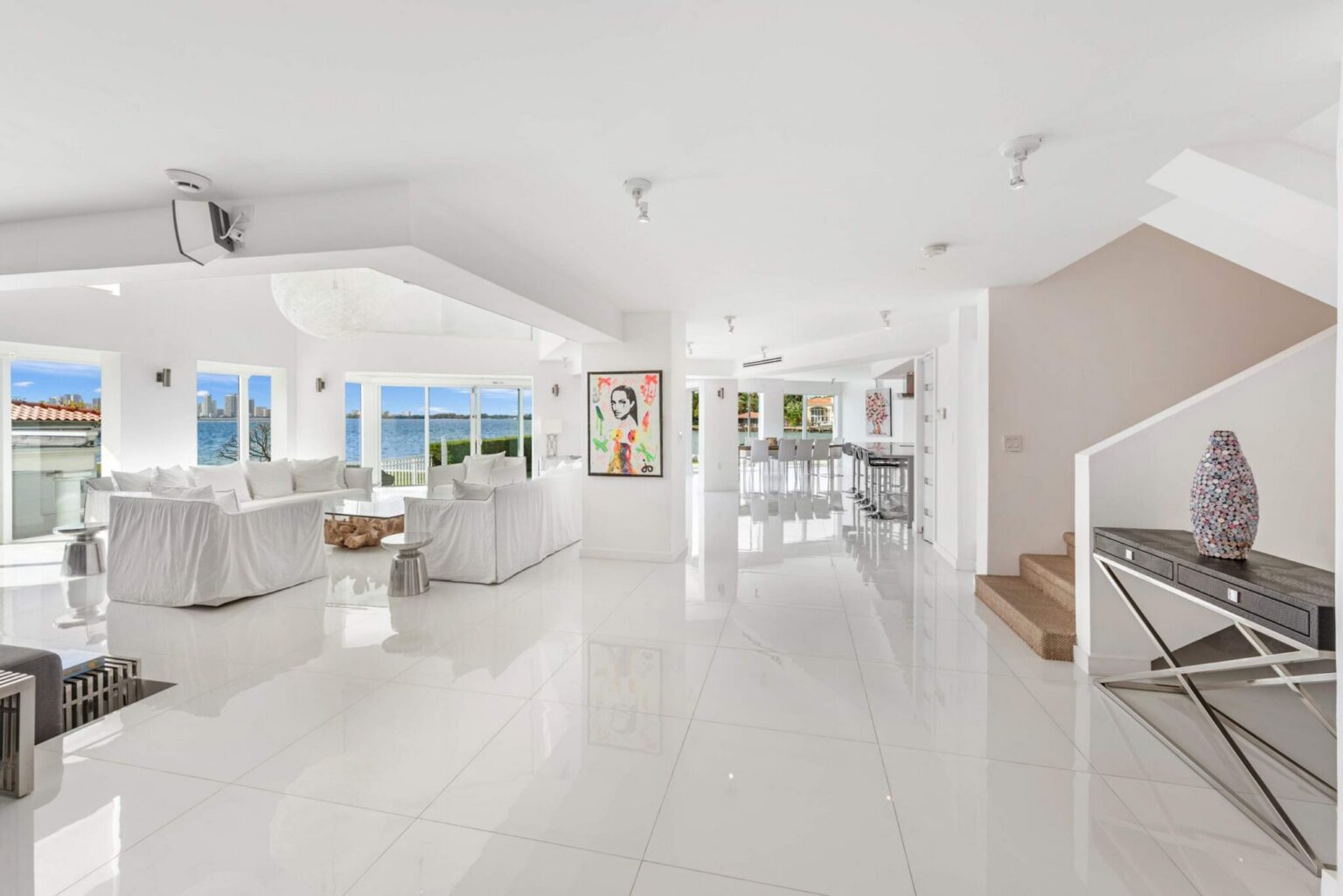 Expansive open-concept main level featuring glossy tile flooring, multiple waterfront seating areas, contemporary artwork, abundant natural light, and seamless flow between the media lounge, kitchen, dining area, and gathering spaces.