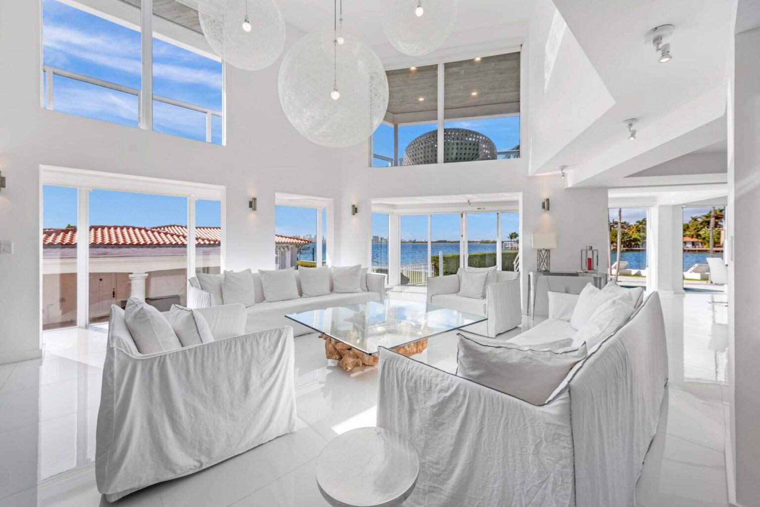 Double-height waterfront living room with vaulted ceilings, floor-to-ceiling windows, oversized lounge seating, designer pendant lighting, glossy tile flooring, and abundant natural light. Open layout connects the kitchen, dining, and living areas.