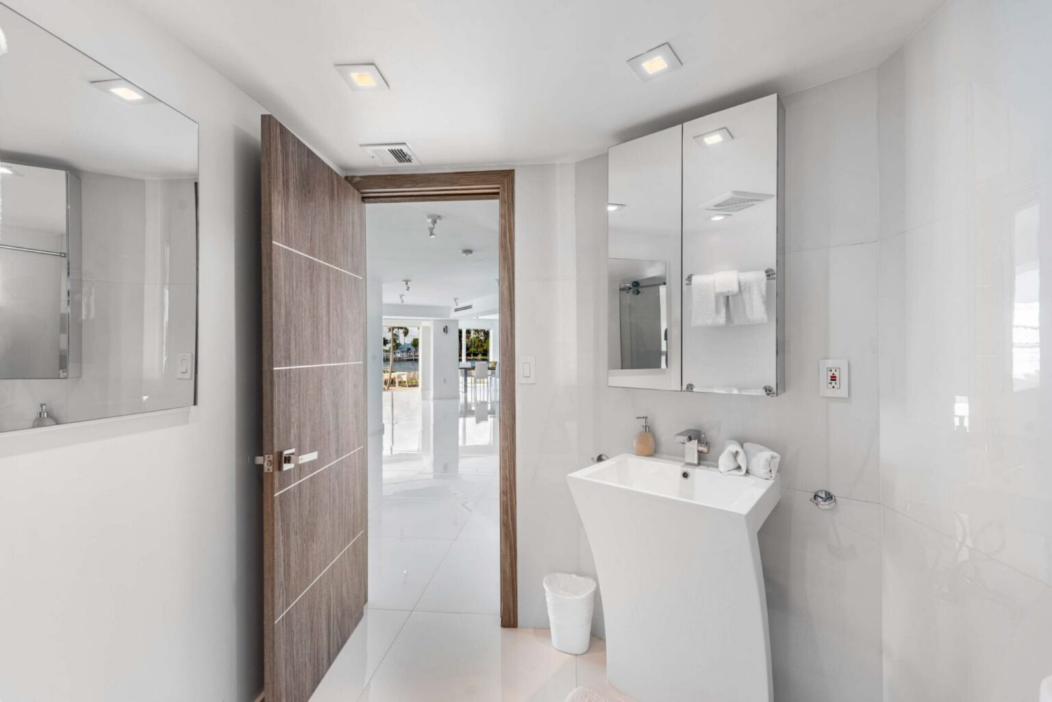 Modern powder room featuring a contemporary pedestal sink, wall-mounted mirror, sleek tile finishes, recessed lighting, and a bright minimalist design. Conveniently located near the main living areas for guest use and accessibility.