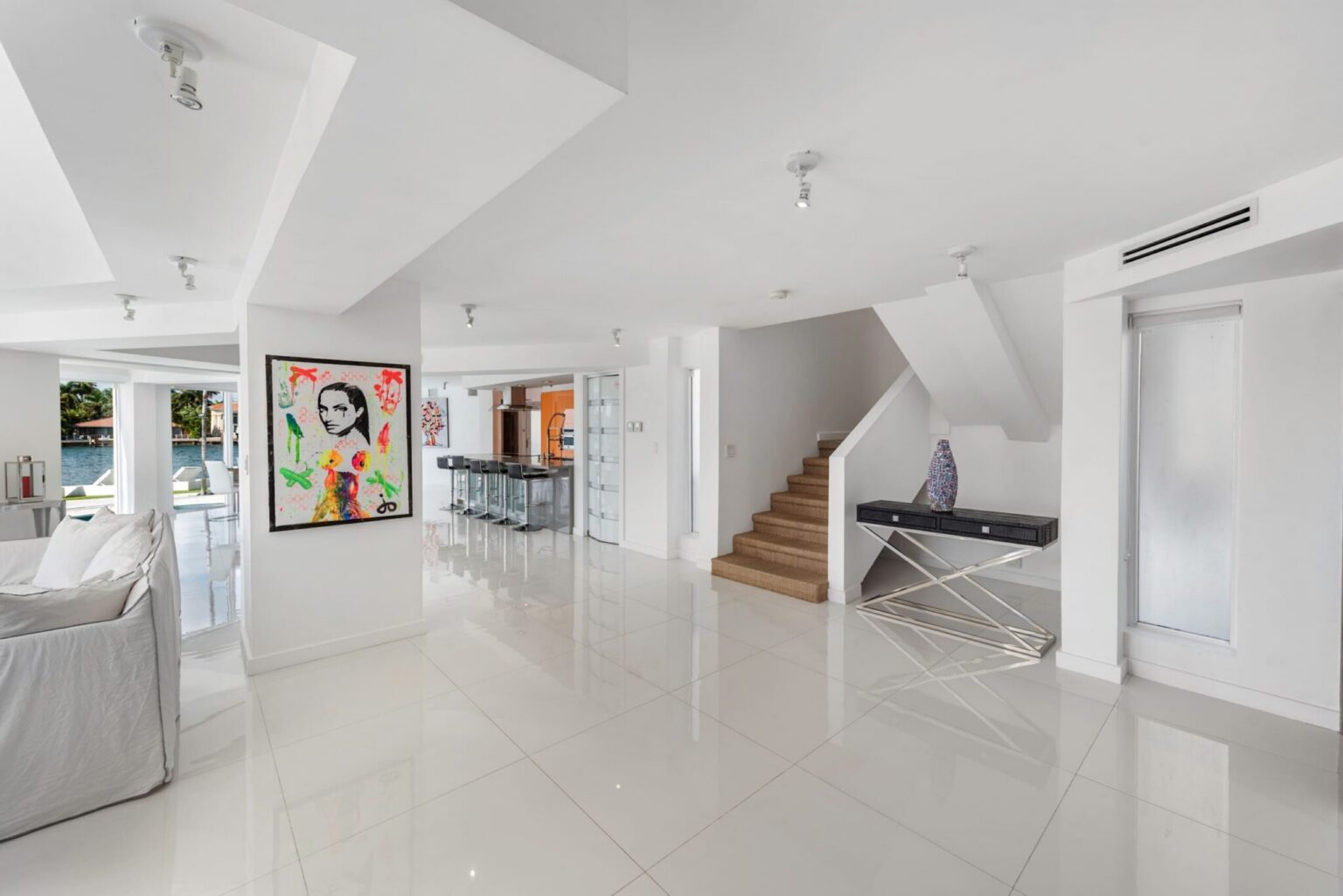 Open foyer featuring glossy tile flooring, contemporary artwork, staircase access, breakfast bar seating, and an open-concept layout. Bright transitional space connects the kitchen, living room, dining area, and guest accommodations.