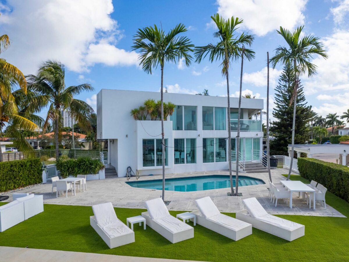 Modern waterfront estate with a private swimming pool, sun loungers, expansive pool deck, mature palm trees, manicured landscaping, floor-to-ceiling windows, and modern architecture overlooking the water.