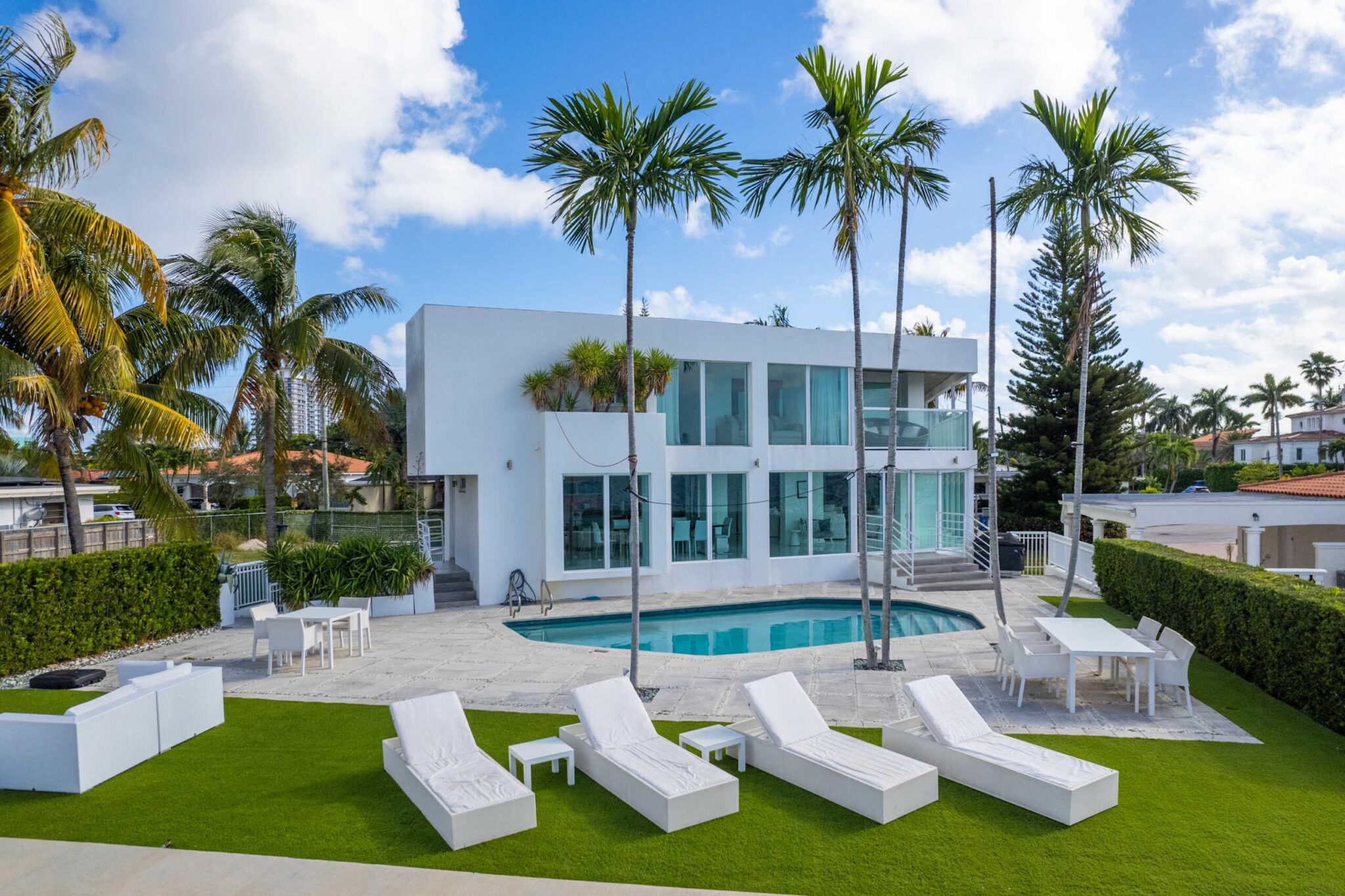 Modern waterfront estate with a private swimming pool, sun loungers, expansive pool deck, mature palm trees, manicured landscaping, floor-to-ceiling windows, and modern architecture overlooking the water.
