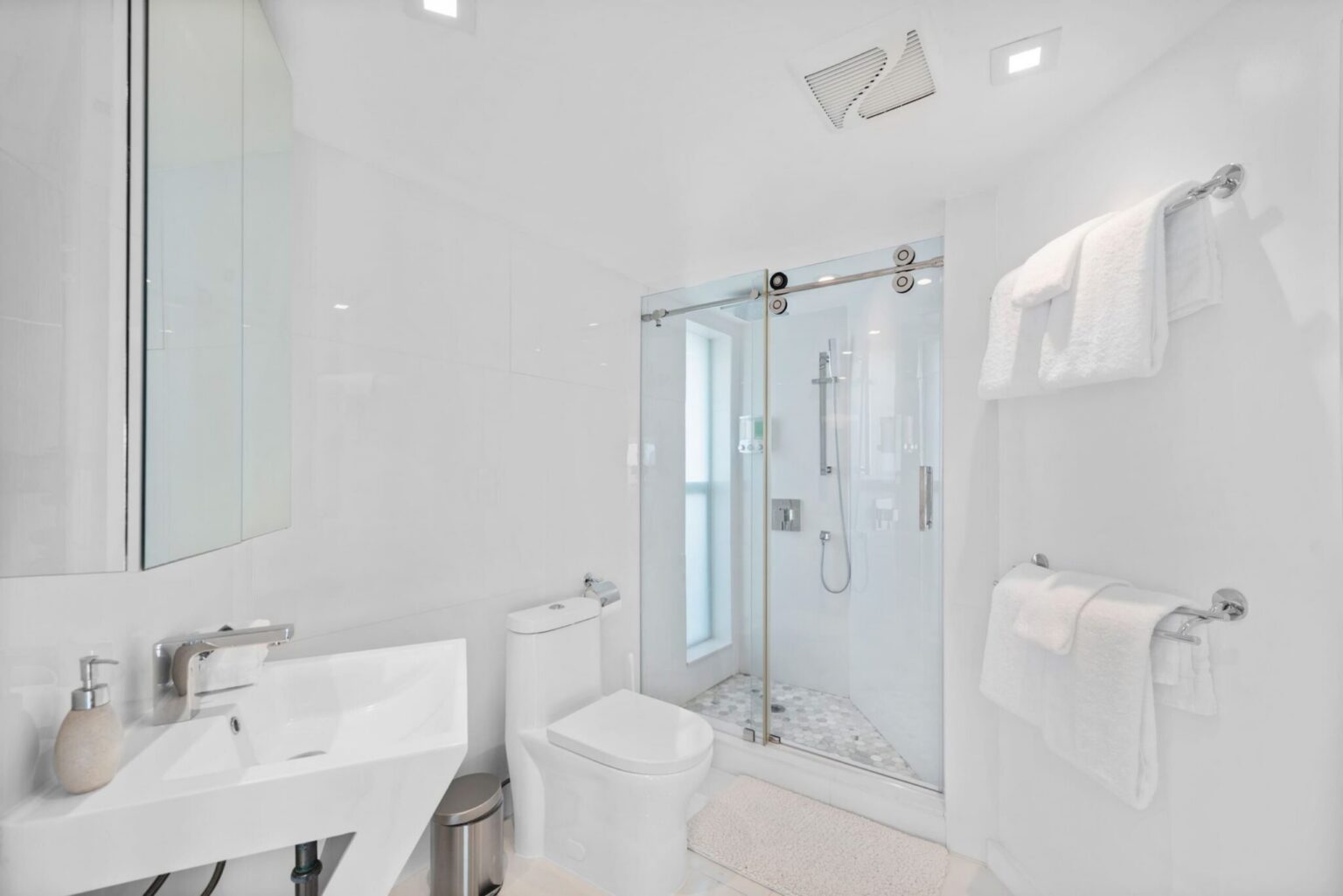 Bright bathroom featuring a glass-enclosed walk-in shower, modern vanity sink, large wall mirror, fresh white tile finishes, recessed lighting, and built-in towel storage. Clean, contemporary layout designed for guest comfort and convenience.