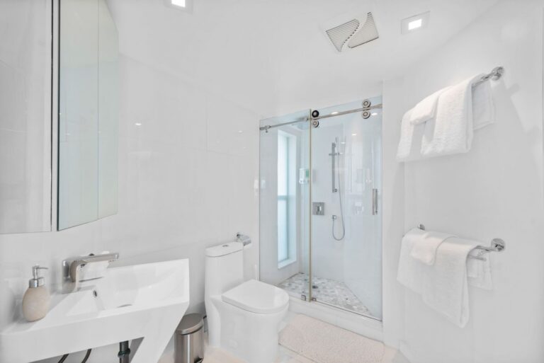 Bright bathroom featuring a glass-enclosed walk-in shower, modern vanity sink, large wall mirror, fresh white tile finishes, recessed lighting, and built-in towel storage. Clean, contemporary layout designed for guest comfort and convenience.