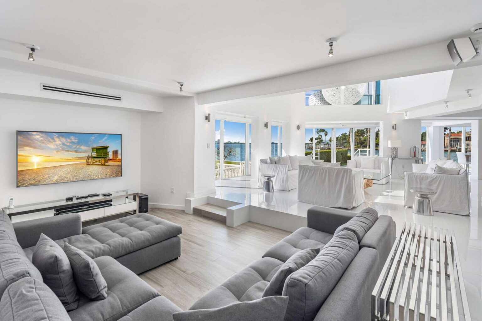 Media lounge featuring an oversized sectional sofa, wall-mounted TV, surround sound speakers, light wood flooring, and large windows. Open-concept layout provides seamless access to the main living room, dining area, and waterfront gathering spaces.