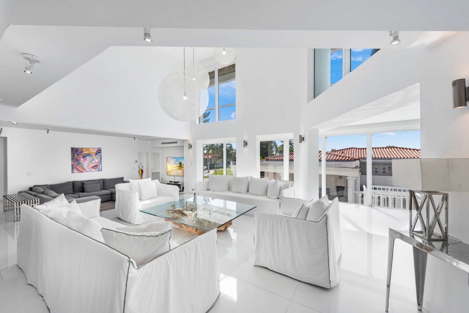 Double-height waterfront living room featuring oversized sectional seating, floor-to-ceiling windows, vaulted ceilings, designer pendant lighting, glossy tile flooring, and an open-concept layout connecting the kitchen, dining, and lounge areas.
