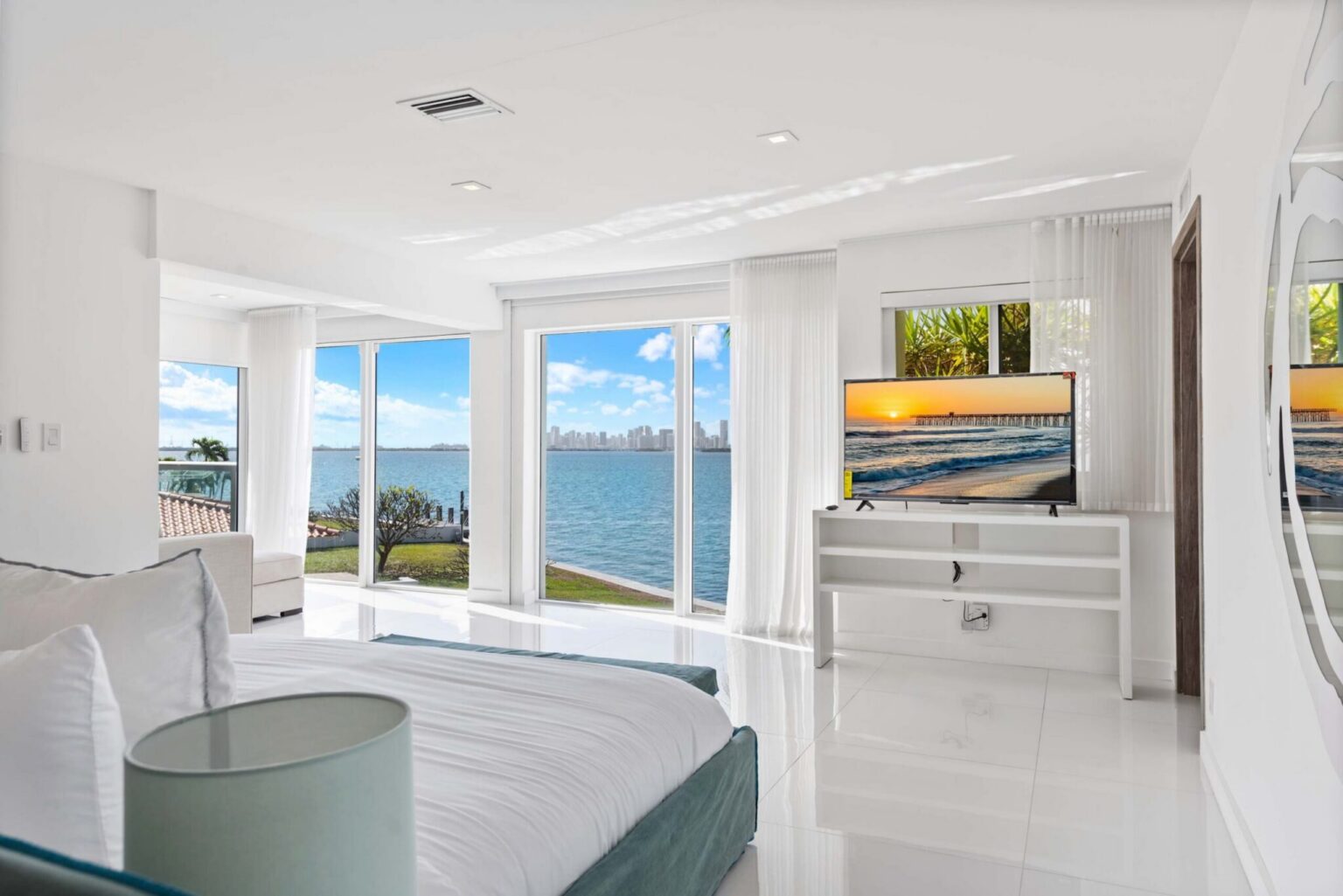 Waterfront primary bedroom featuring a king-size bed, floor-to-ceiling windows, panoramic bay views, white tile flooring, wall-mounted TV, modern furnishings, and abundant natural light throughout the spacious open layout.