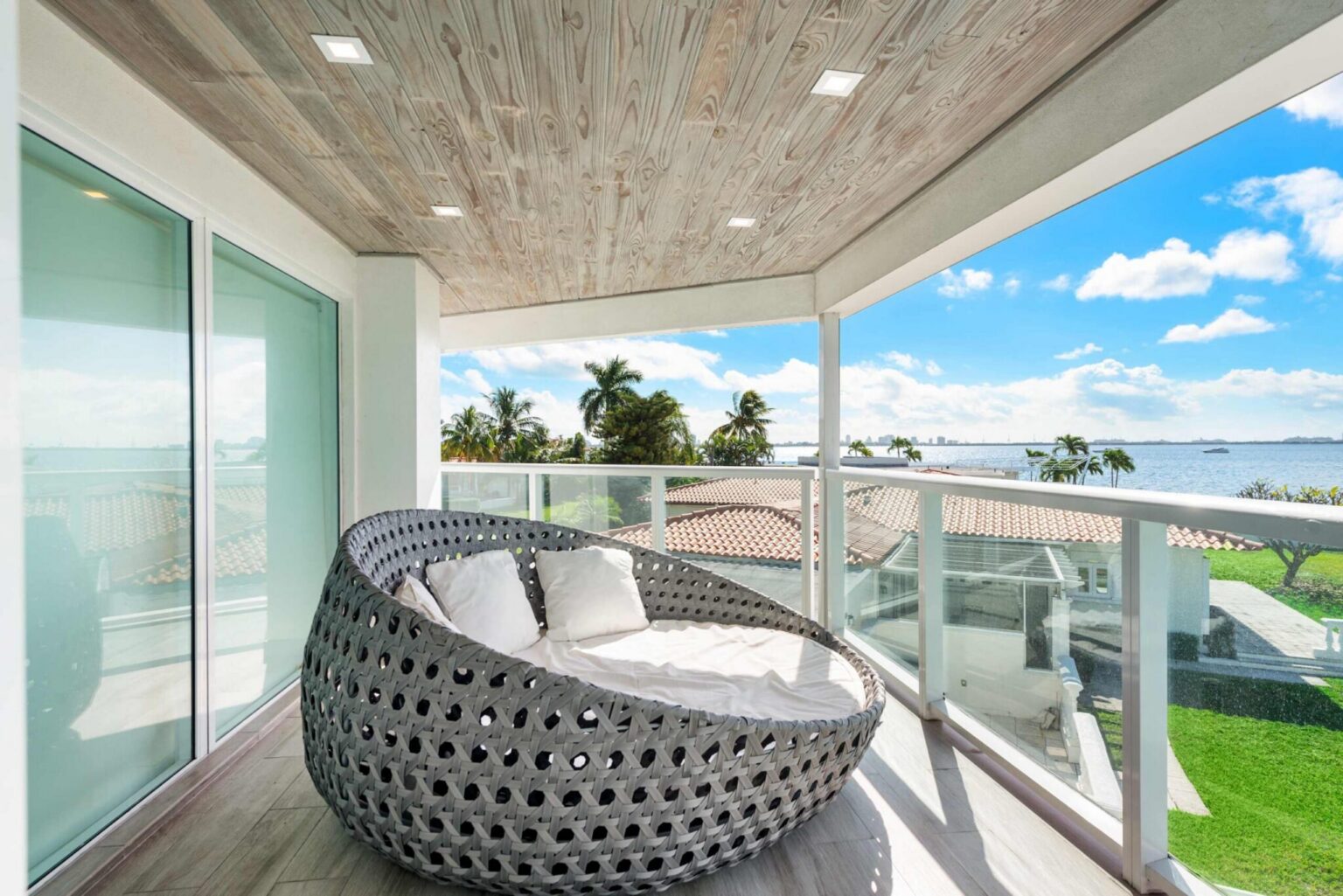 Covered upper-level balcony featuring a woven outdoor daybed, glass railings, wood-accent ceiling, and elevated views of the waterfront and surrounding neighborhood. Private outdoor seating area with direct access from the interior living spaces.