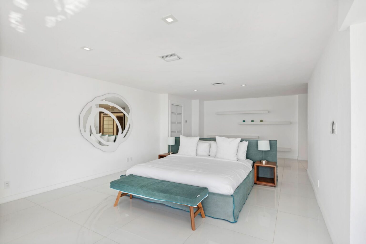 Spacious bedroom featuring a king-size bed, modern furnishings, wall-mounted reading lights, minimalist décor, white tile flooring, bedside tables, and ample natural light. Clean, contemporary design with an open and airy layout.