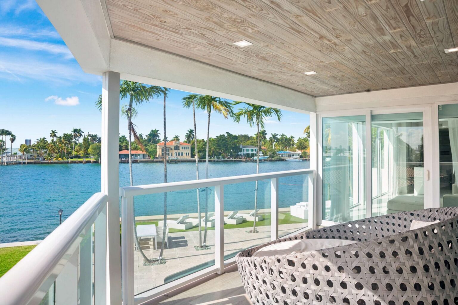 Covered waterfront balcony featuring outdoor lounge seating, glass railings, wood-accent ceiling, panoramic water views, and direct access from the interior living spaces. Ideal for enjoying the scenery and outdoor relaxation areas.