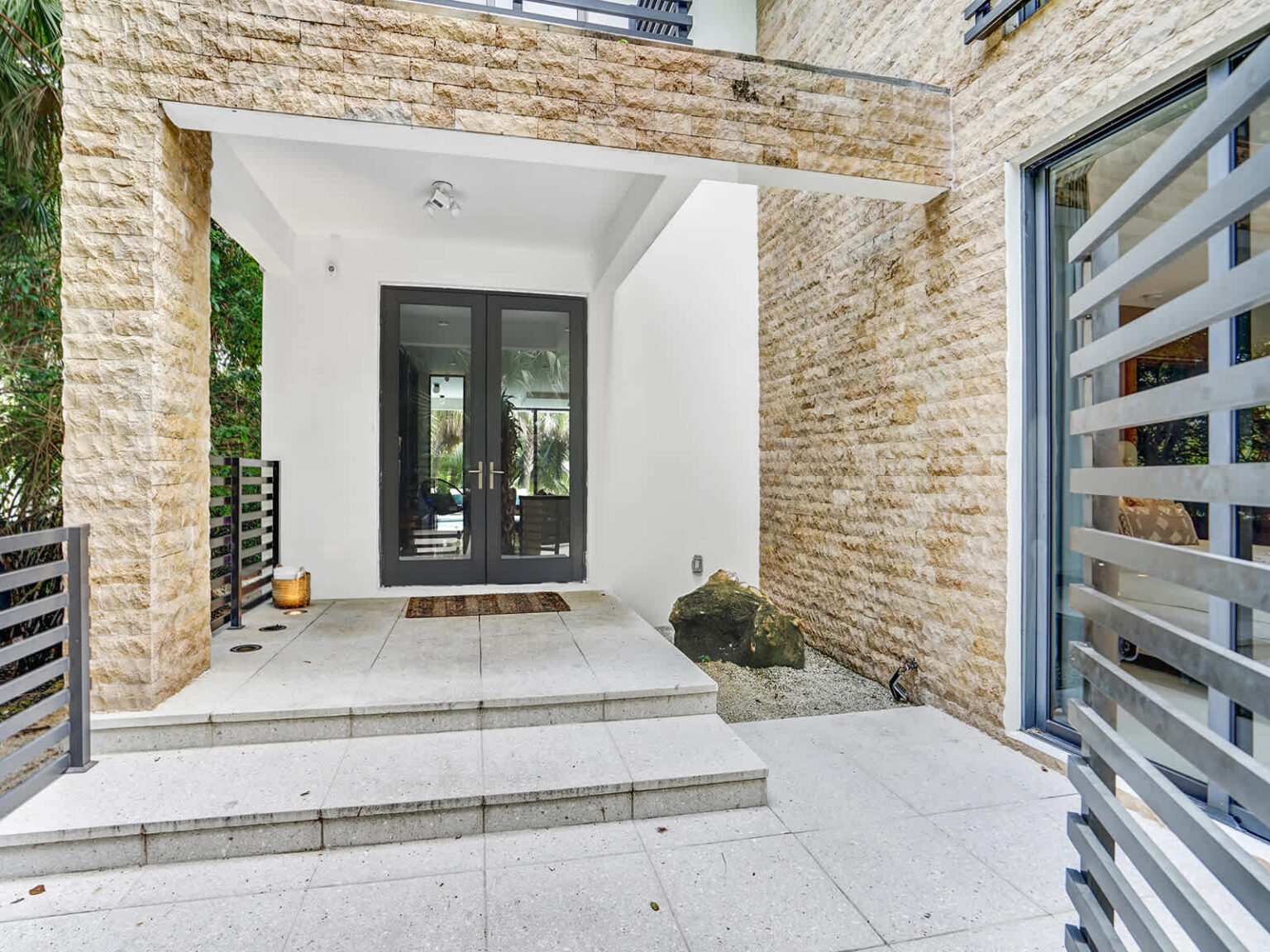 Covered front entryway featuring modern architecture, natural stone accent walls, and double glass entry doors. The spacious tiled porch provides a welcoming transition into the home, with clean lines and contemporary finishes throughout.
