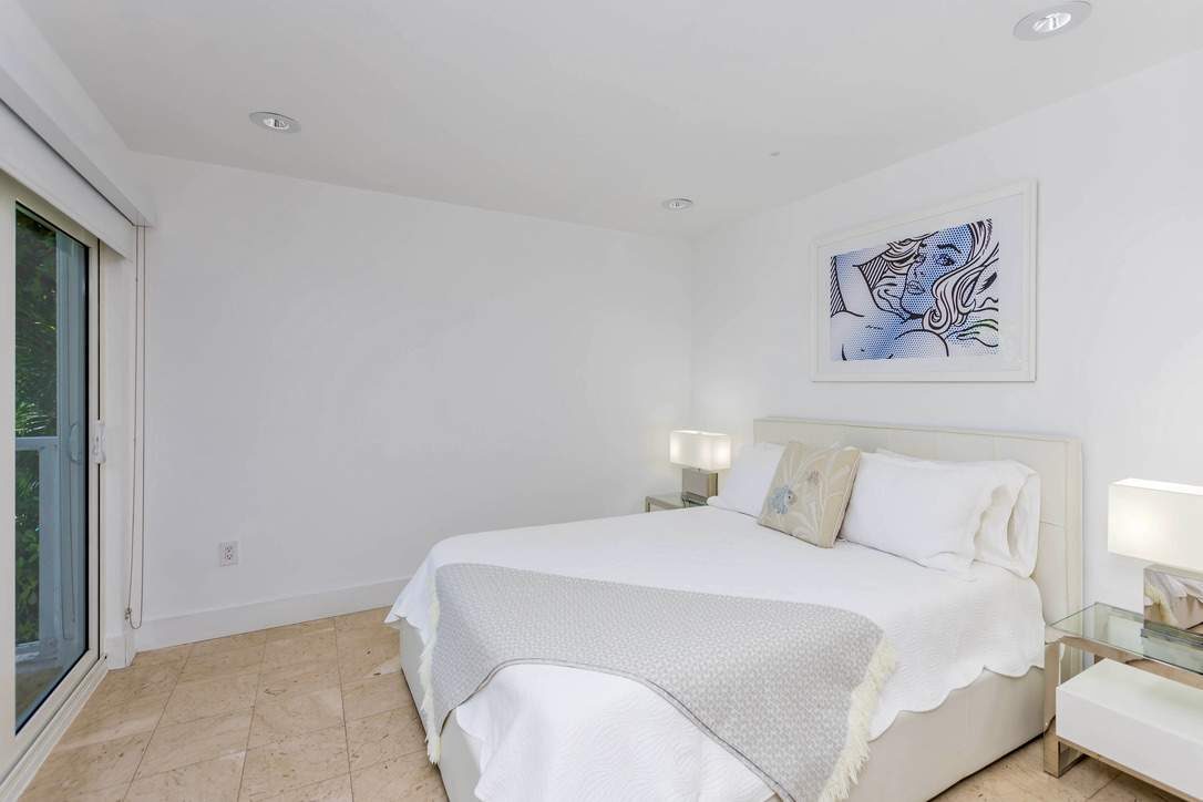 Bright guest bedroom featuring a queen-size bed with white linens, bedside tables with lamps, tile flooring, contemporary wall art, recessed lighting, and sliding glass doors providing natural light and convenient outdoor access.