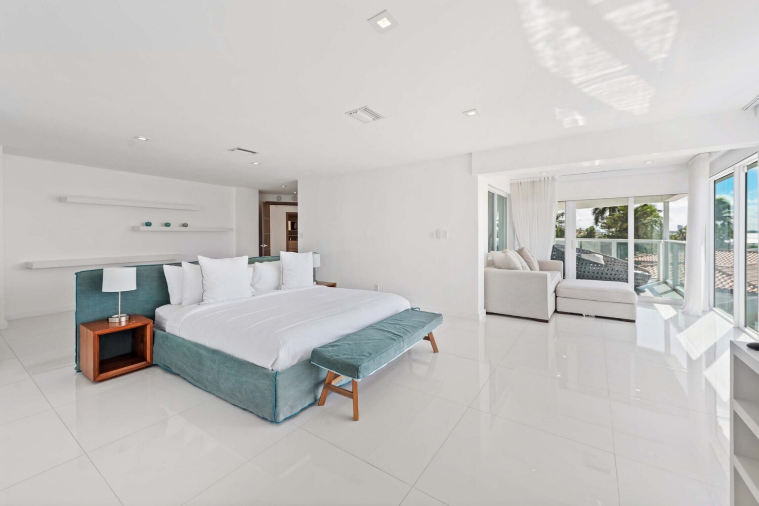 Expansive primary suite featuring a king-size bed with premium linens, bench seating, tile flooring, bedside tables with lamps, a private sitting area, floor-to-ceiling windows, and an open layout filled with natural light.