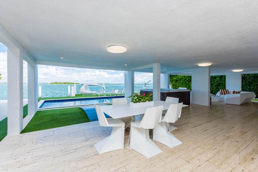 Covered outdoor dining and lounge area featuring a dining table with seating for six, open-air waterfront views, wood-look tile flooring, landscaped accents, built-in entertaining space, and direct access to the private pool terrace and outdoor ameni