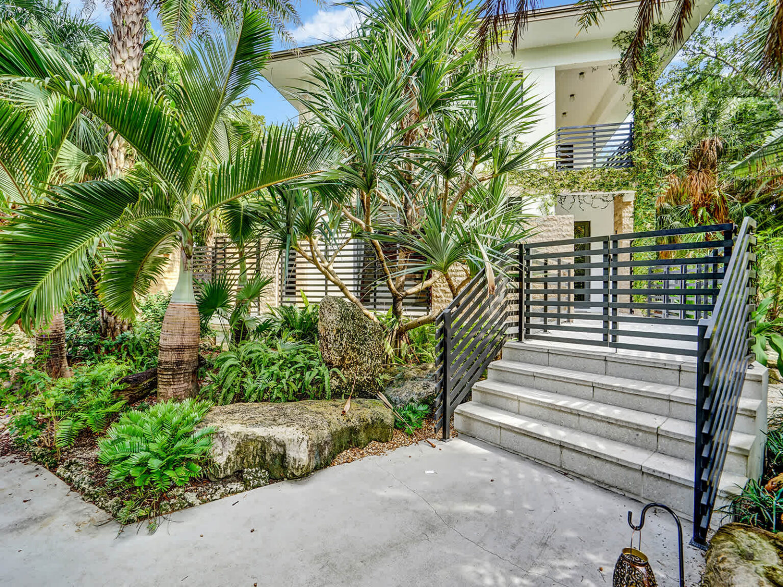 Landscaped entryway featuring tropical gardens, mature palm trees, and natural stone accents. Exterior stairs with modern railings lead to the home, while lush greenery and shaded surroundings create a welcoming arrival experience.