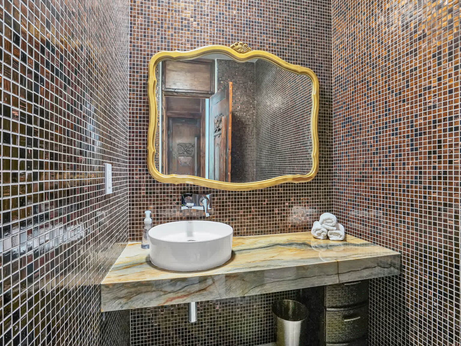 Contemporary powder room featuring a vessel sink, stone countertop, and decorative mosaic tile walls. An illuminated framed mirror adds character, while modern fixtures and natural finishes create a clean, functional design.