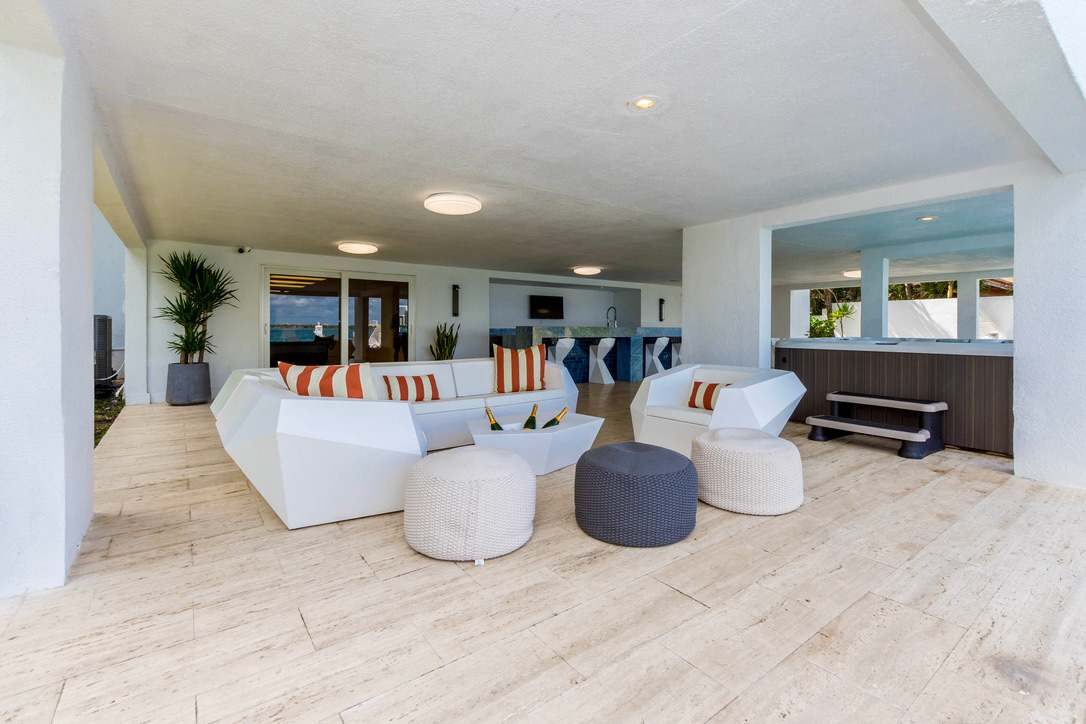 Poolside entertainment lounge featuring contemporary outdoor sofas, cushioned seating, accent ottomans, a custom stone bar, integrated spa area, wood-look tile flooring, recessed lighting, and a covered open-air layout designed for gathering and rela