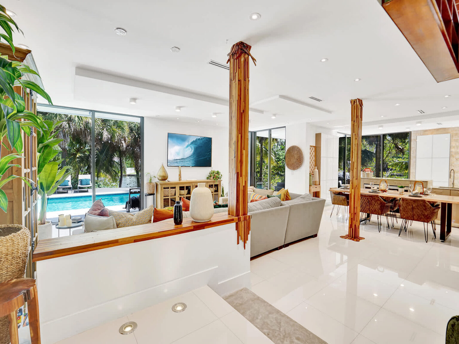Open living room with large picture windows overlooking the pool and tropical landscaping. Features high ceilings, polished tile flooring, modern seating, natural wood columns, and seamless indoor-outdoor connectivity.