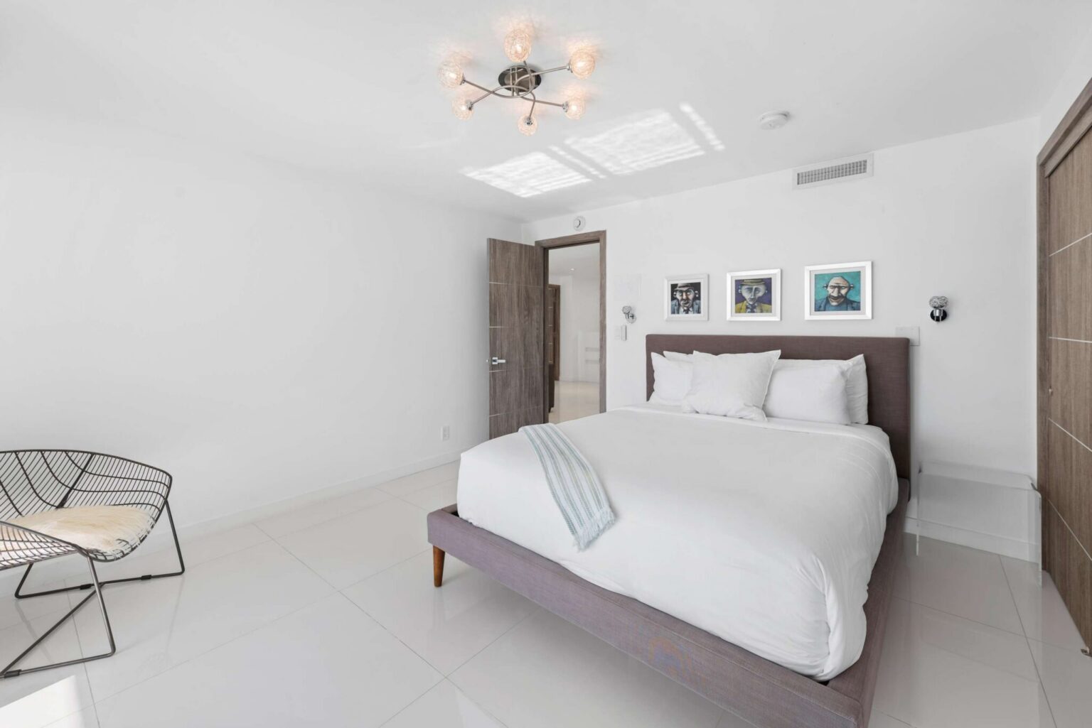 Modern guest bedroom featuring a queen-size bed with white linens, tile flooring, contemporary ceiling lighting, bedside reading lamps, framed artwork, and a spacious minimalist layout designed for comfort, relaxation, and extended stays.