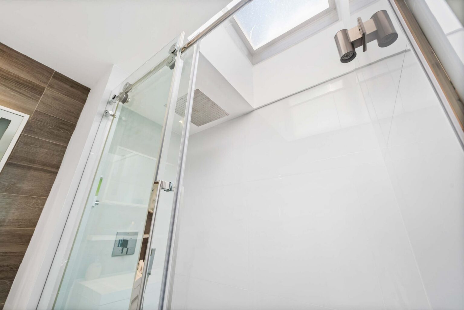 Walk-in shower enclosure featuring a frameless glass door, modern chrome fixtures, skylight for natural light, recessed lighting, and clean contemporary finishes designed to enhance brightness, ventilation, and functionality within the bathroom space