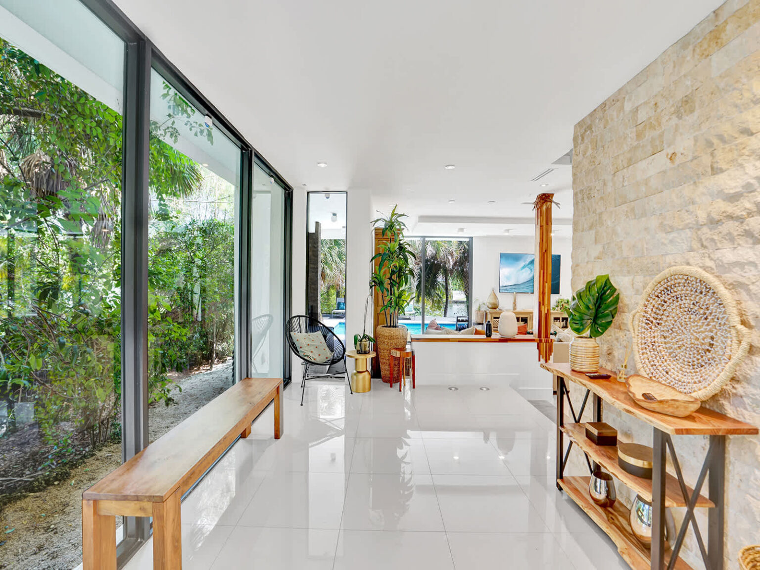 Bright open-concept living area with floor-to-ceiling windows, polished tile flooring, and abundant natural light. Features modern furnishings, indoor plants, and seamless views of the outdoor patio, pool, and tropical landscaping.
