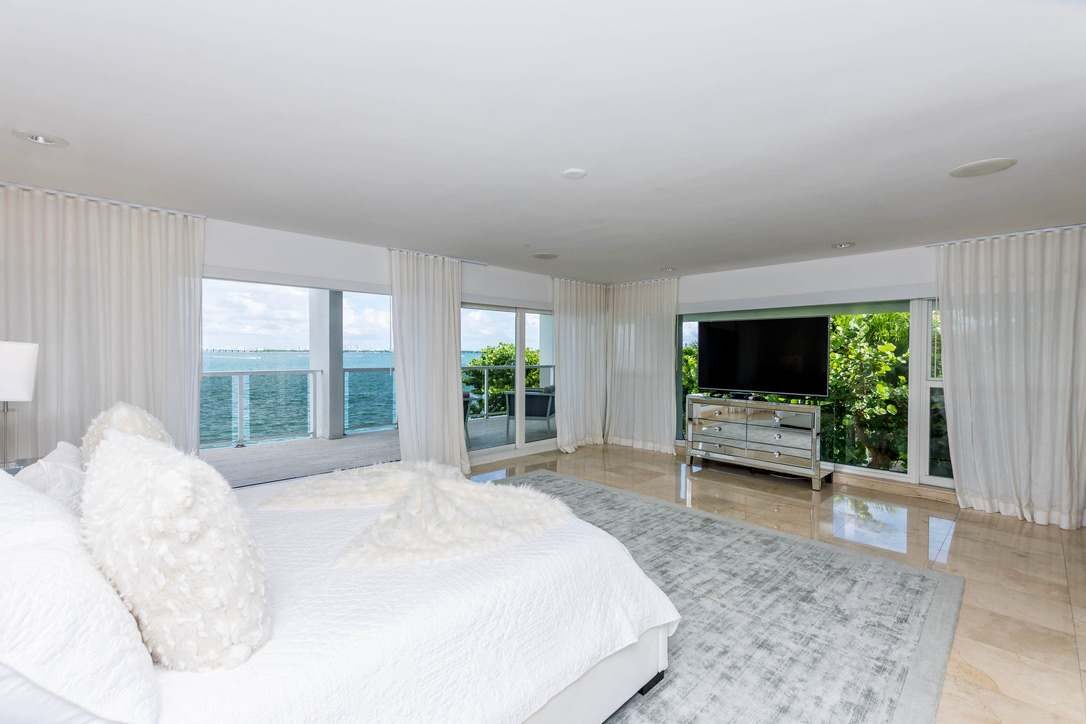 Panoramic primary suite featuring a king-size bed with premium linens, floor-to-ceiling windows, wall-mounted smart TV, oversized area rug, tile flooring, and expansive water views with direct access to a private waterfront balcony.