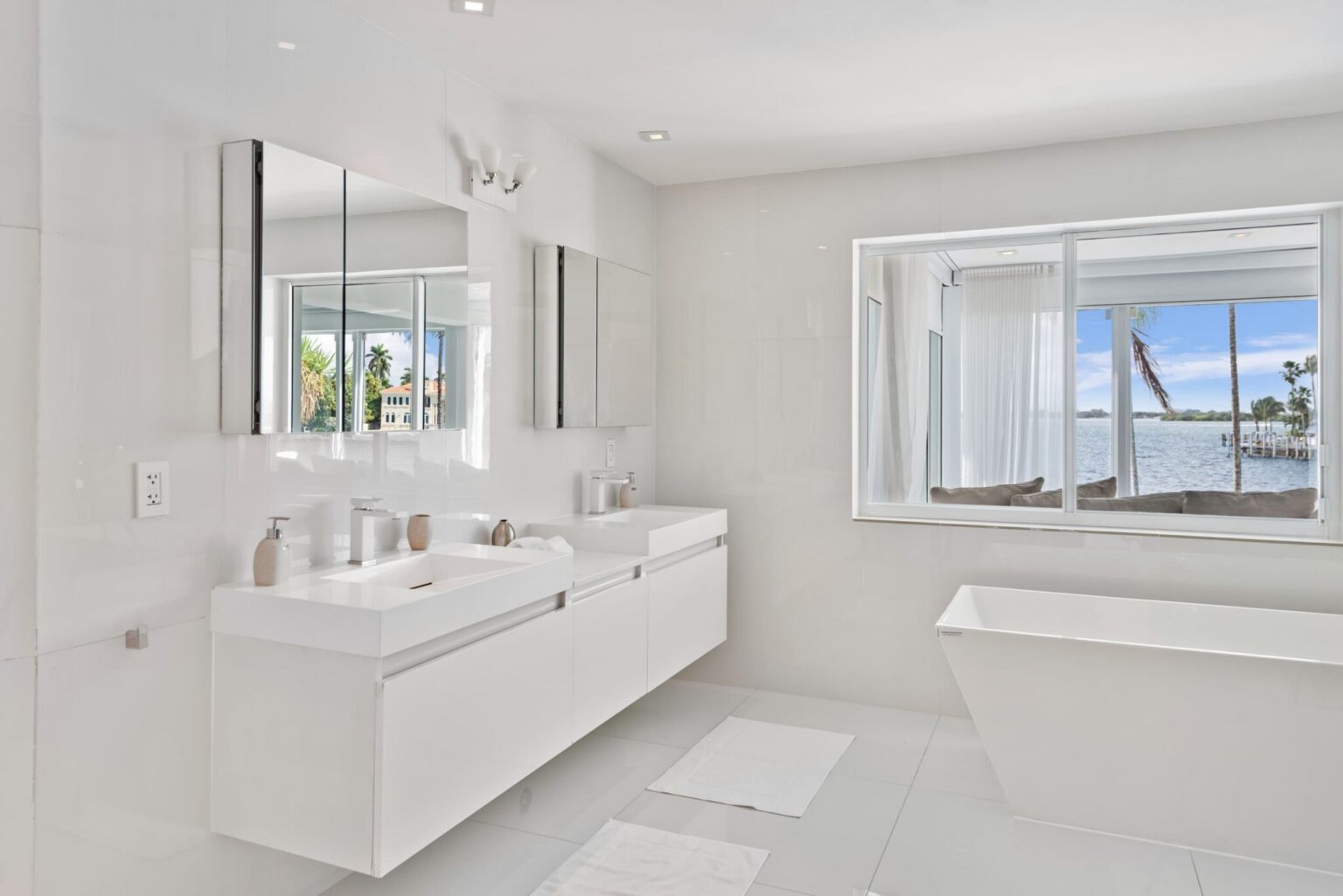 Bright primary bathroom featuring a double-sink floating vanity, freestanding soaking tub, oversized mirror, tile flooring, and large windows with water views. Clean contemporary design offers ample counter space and natural light.