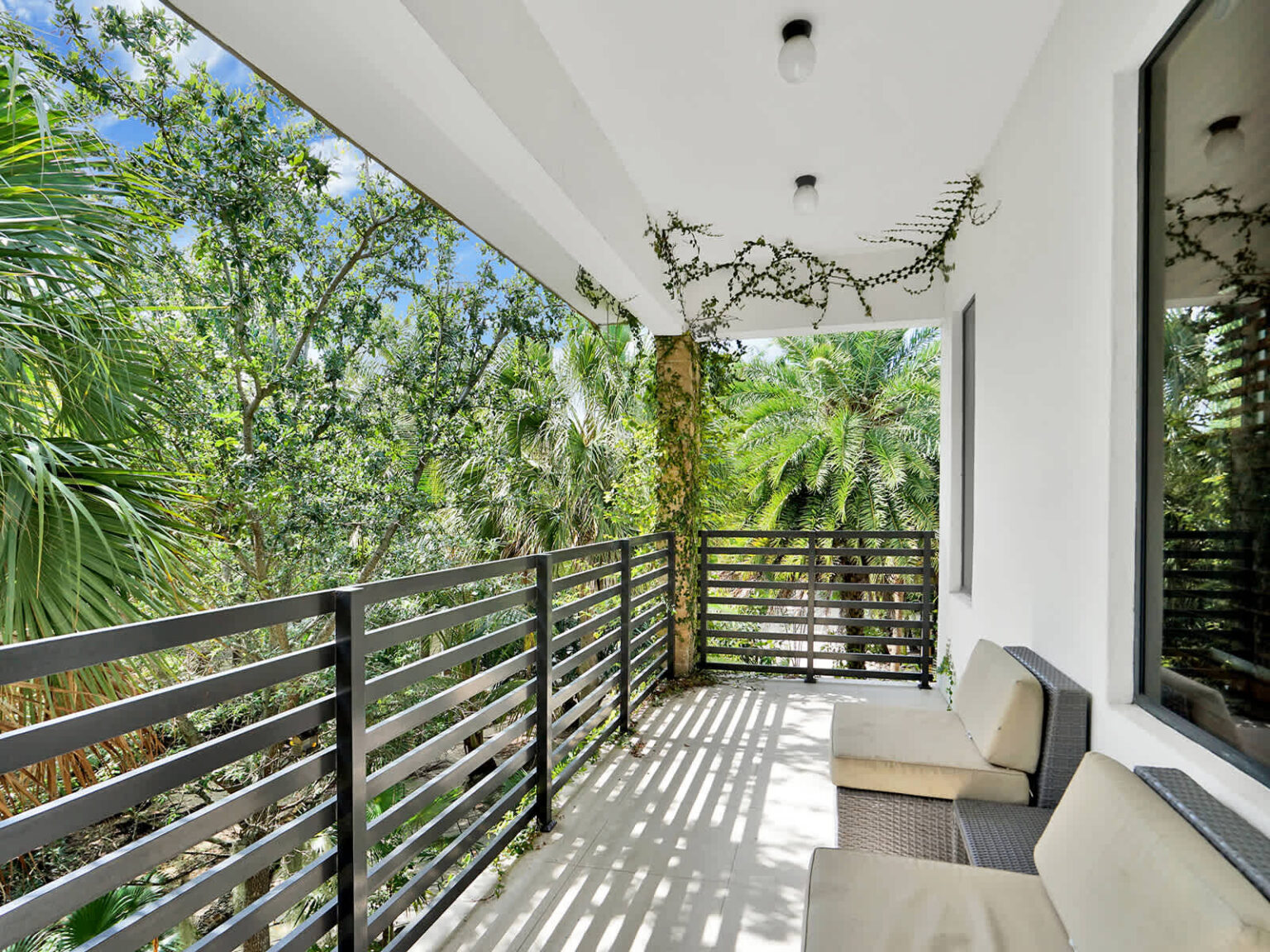 Covered balcony with modern metal railings, outdoor seating, and views of lush tropical greenery. This shaded outdoor space extends the living area, providing a comfortable setting for relaxation, fresh air, and garden views.