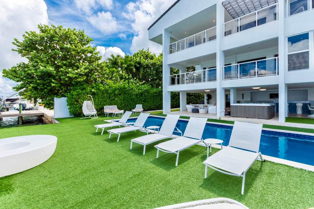 Waterfront villa outdoor area with a private swimming pool, sun loungers, artificial turf lawn, landscaped tropical greenery, covered patio space, multiple balconies, and direct access to poolside relaxation and entertainment areas.