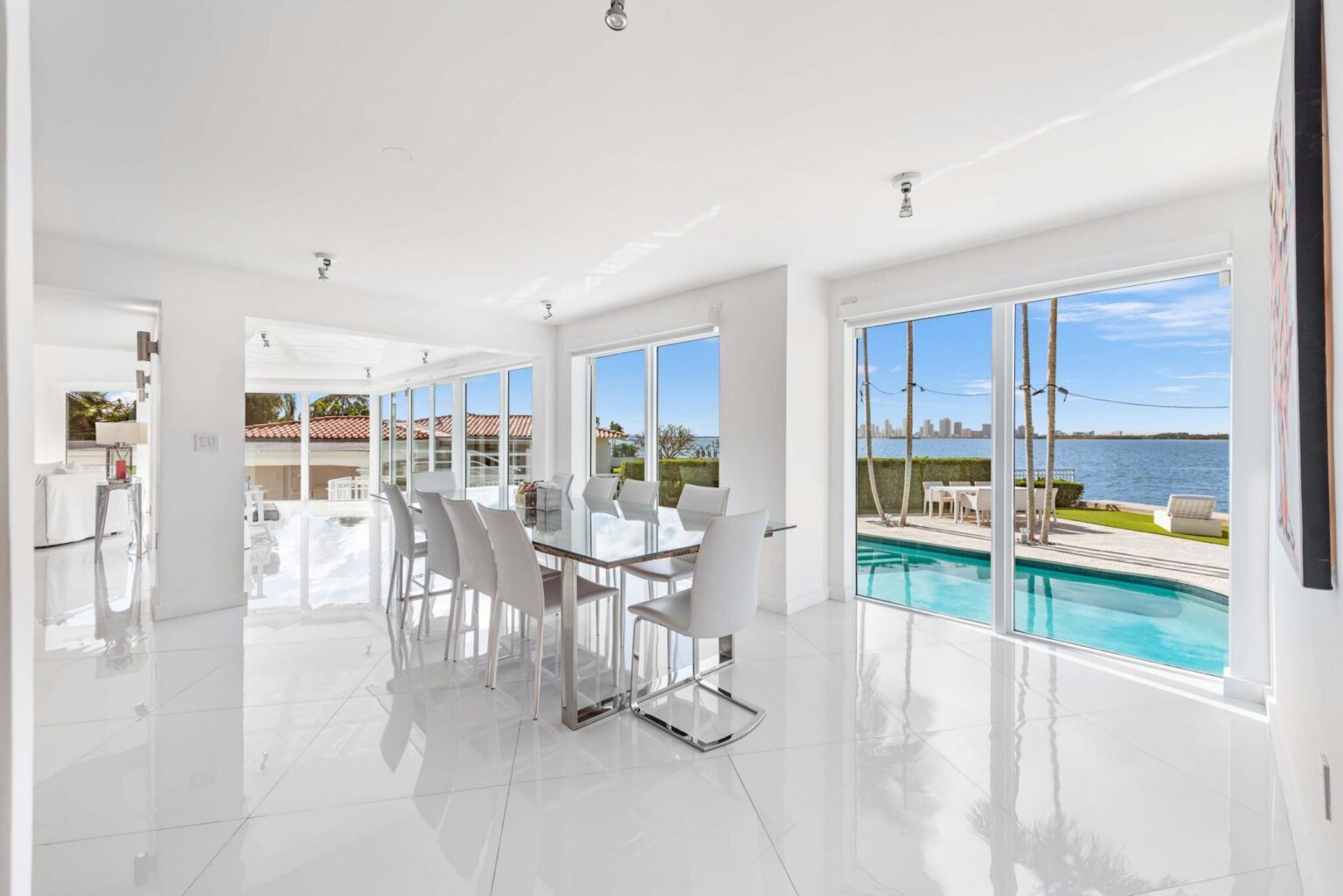 Waterfront dining space featuring a contemporary dining table with seating for six, floor-to-ceiling windows, tile flooring, and panoramic views of the private pool, bay, and city skyline. Bright open layout connects seamlessly to the kitchen.