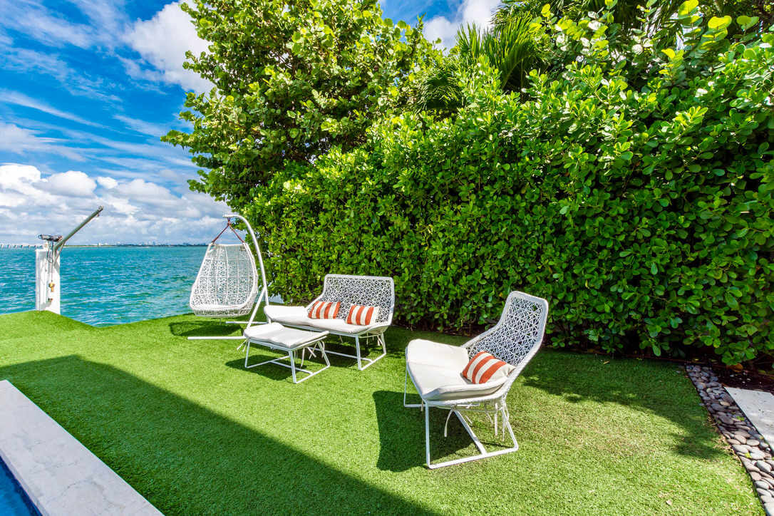 Outdoor garden seating area featuring cushioned lounge chairs, a hanging egg chair, artificial turf lawn, mature privacy hedges, and direct waterfront access. Located beside the pool terrace with bay views and dedicated outdoor relaxation space.