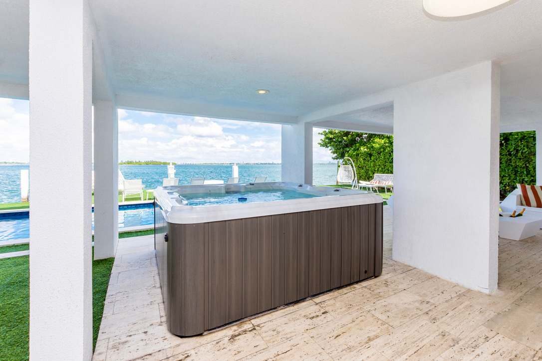 Covered outdoor spa area featuring a built-in hot tub, open-air waterfront views, wood-look tile flooring, landscaped privacy hedges, shaded lounge space, and direct access to the pool terrace, outdoor dining area, and waterfront amenities.