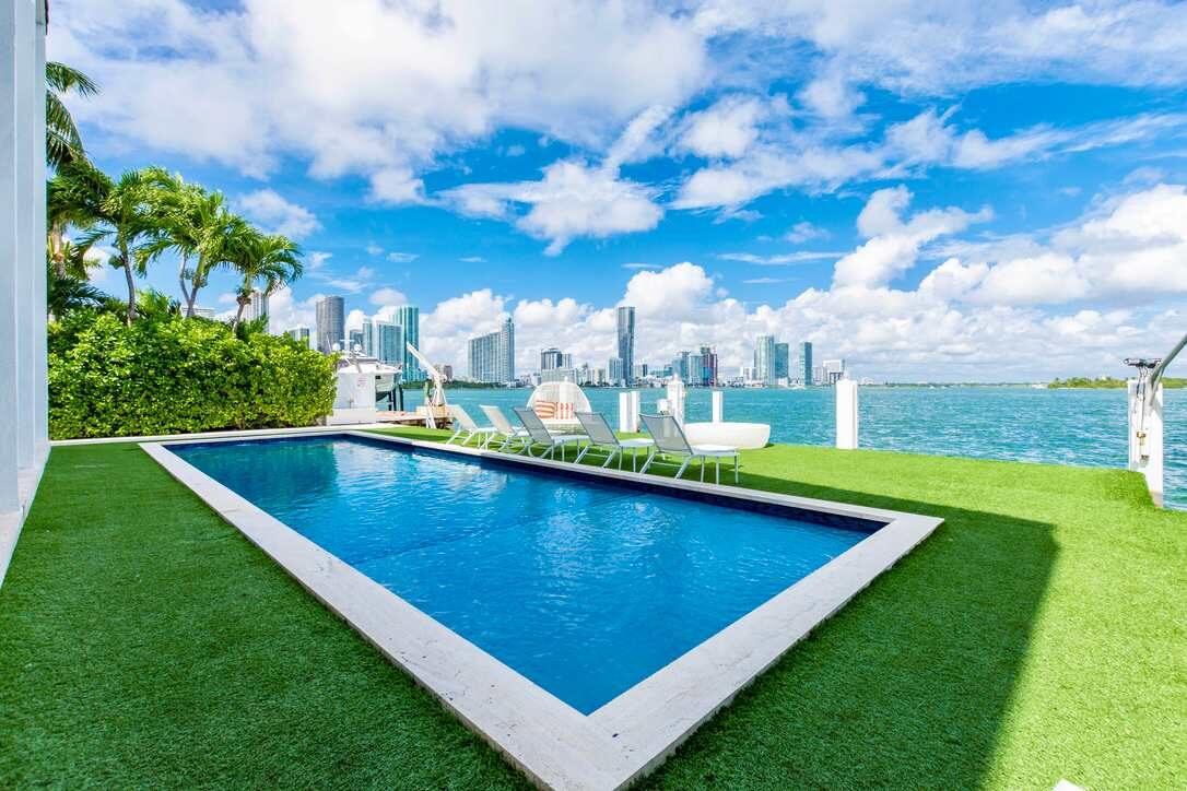 Waterfront pool area featuring a private swimming pool, artificial turf lawn, poolside sun loungers, glass safety fencing, landscaped greenery, and unobstructed bay and city skyline views from the outdoor terrace.