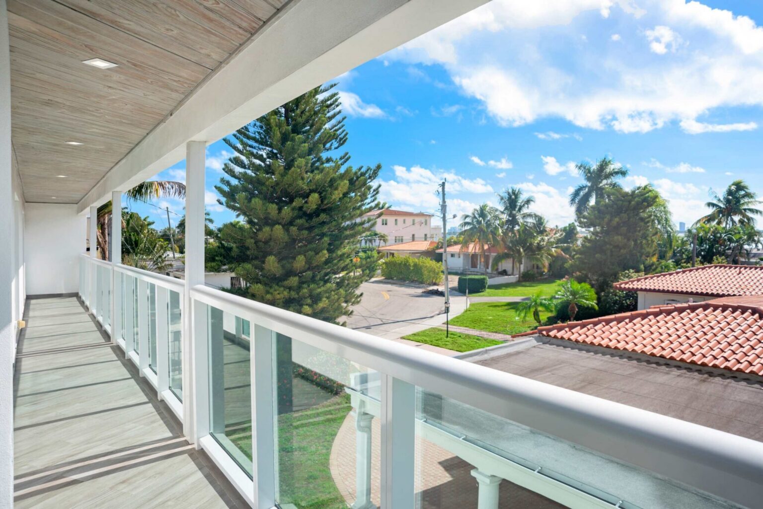 Elevated balcony featuring glass railings, covered outdoor space, wood look flooring, and scenic views of tropical landscaping and neighboring waterfront homes. Provides convenient access between upper-level rooms while maximizing natural light.