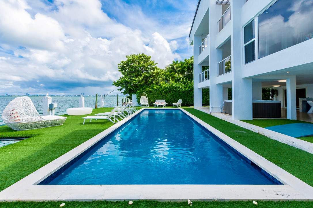Waterfront outdoor area featuring a private lap pool, artificial turf lawn, poolside seating, glass safety fencing, covered patio space, and panoramic water views. Located alongside a modern multi-story villa with direct access to outdoor amenities.