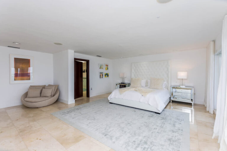 Oversized primary bedroom featuring a king-size bed with upholstered headboard, bedside tables, lounge chair, area rug, tile flooring, recessed lighting, wall art, air conditioning, and a spacious open-plan layout with natural light.