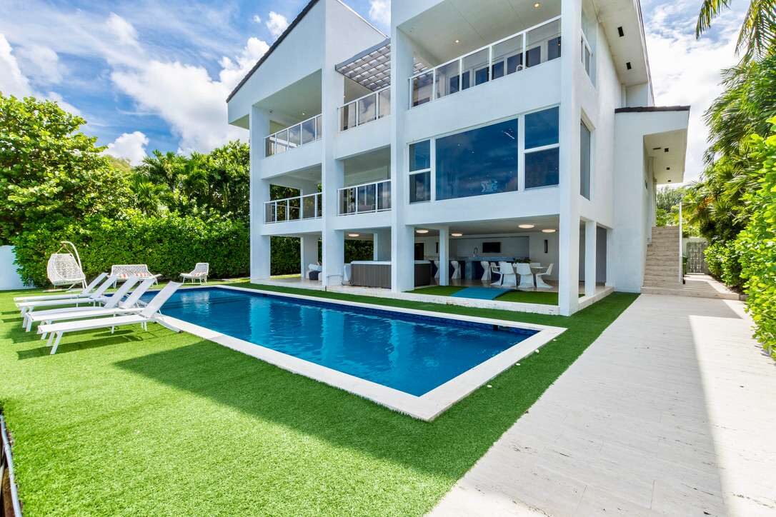 Modern villa exterior featuring a private swimming pool, poolside sun loungers, expansive artificial turf lawn, covered outdoor entertainment area, multiple balconies, floor-to-ceiling windows, landscaped privacy hedges, and contemporary architecture