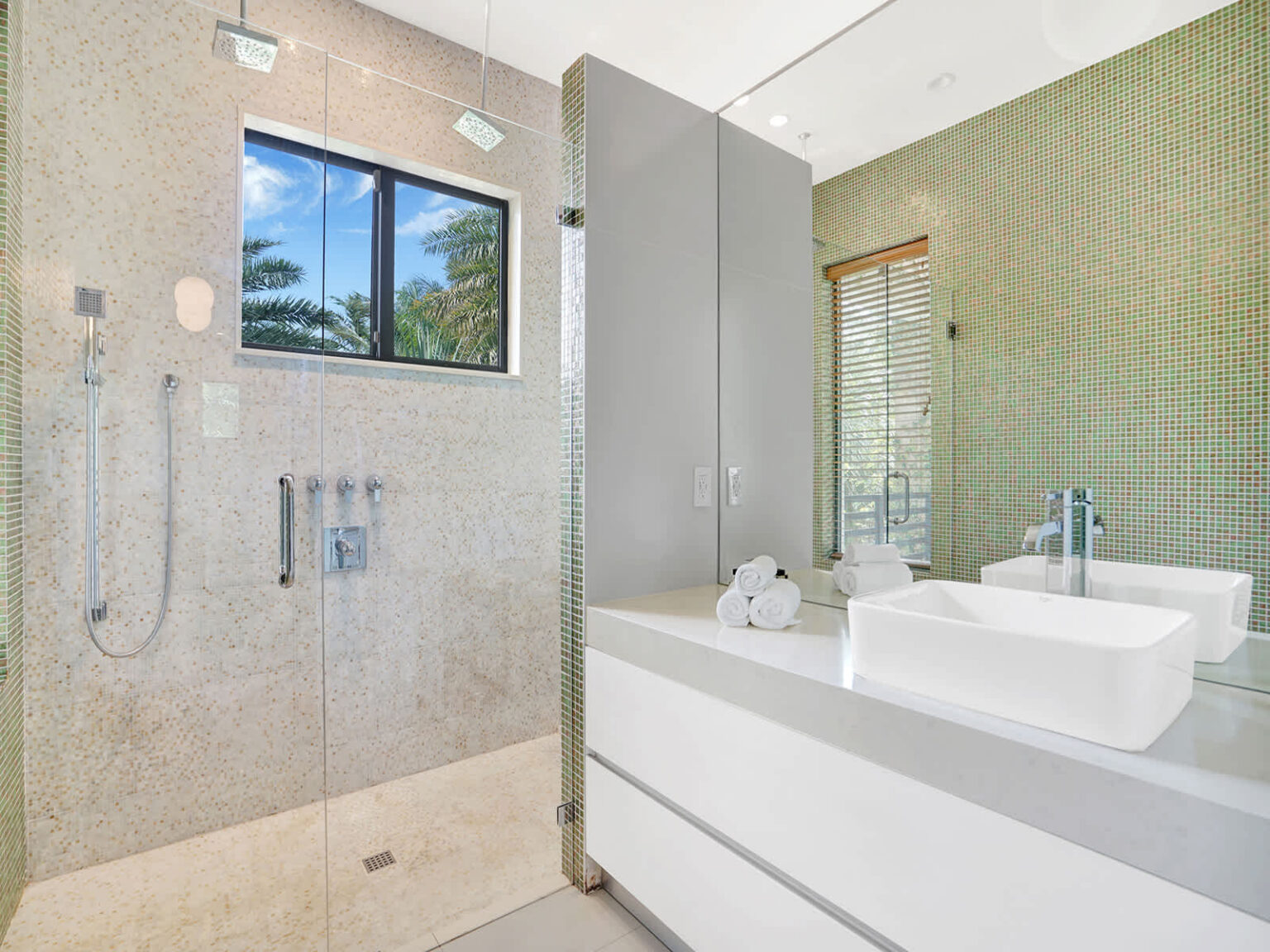 Contemporary bathroom featuring a walk-in glass shower, modern vanity with vessel sink, and natural stone finishes. A large window provides natural light and tropical views, creating a bright, functional, and well-appointed space.