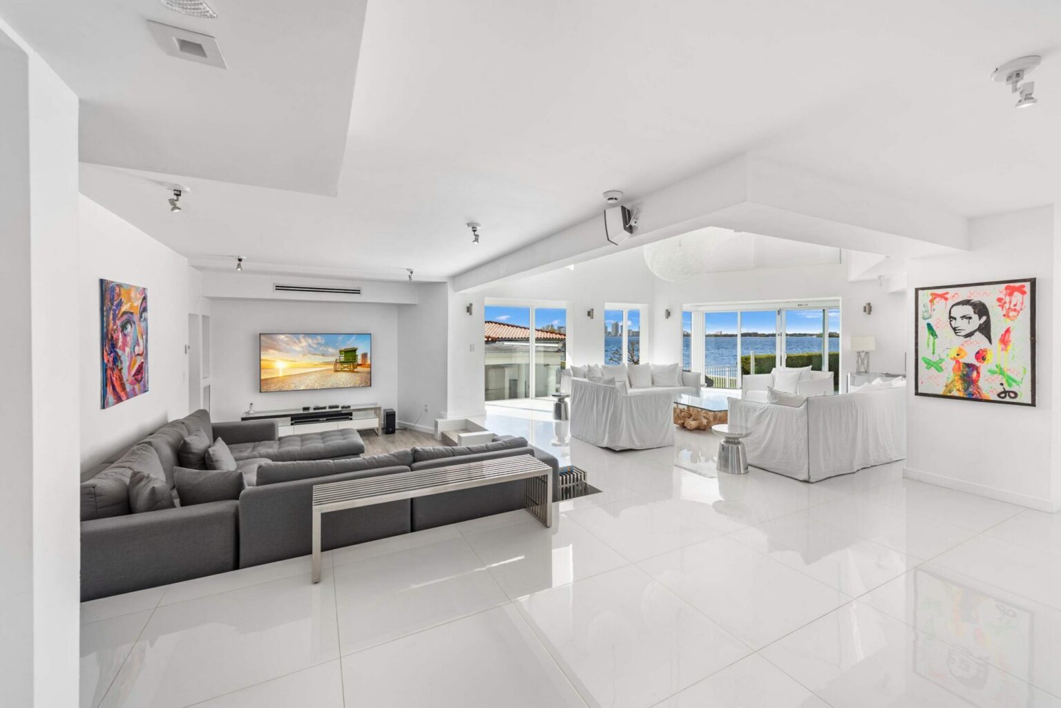 Open-concept entertainment and living area featuring a media lounge with oversized sectional seating, wall-mounted TV, glossy tile flooring, contemporary artwork, expansive windows, and seamless flow to the waterfront living spaces.