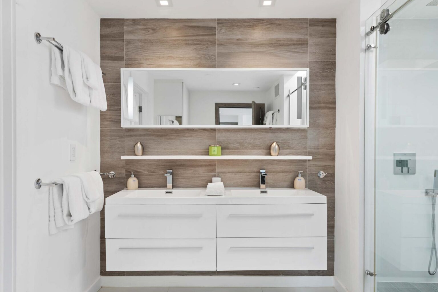 Designer bathroom vanity area featuring a double-sink floating vanity, oversized illuminated wall mirror, wood-look accent tile, modern fixtures, towel storage, and a glass-enclosed walk-in shower within a bright contemporary layout.