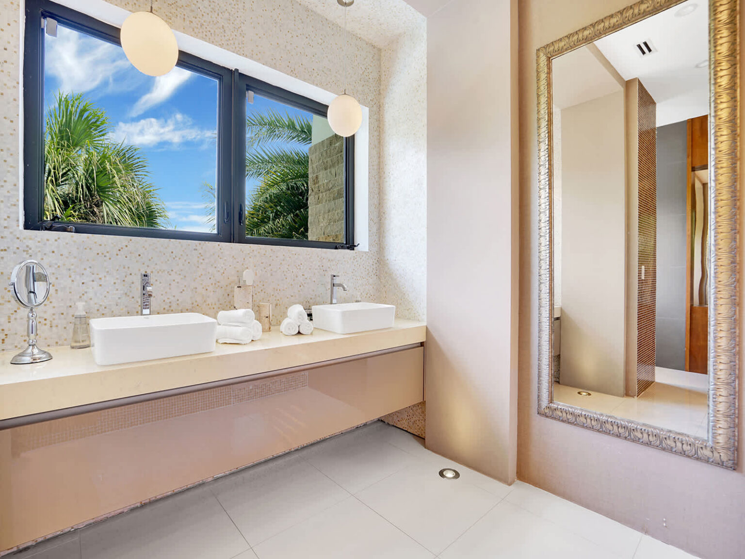 Contemporary bathroom featuring a soaking tub beneath a large picture window, modern vanity, and oversized framed mirror. Natural light, clean finishes, and tropical views create a bright, functional, and relaxing space.