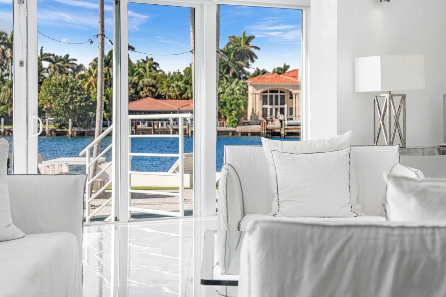 Waterfront lounge corner featuring plush armchair seating, floor-to-ceiling glass doors, tile flooring, and views of the bay and neighboring waterfront homes. Bright sitting area provides direct access to outdoor spaces and natural light.