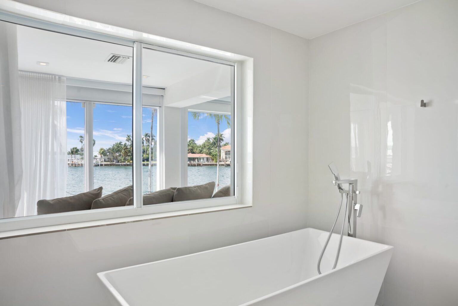 Freestanding soaking tub positioned beside a large interior window, featuring tile flooring, contemporary fixtures, bright natural light, and views into the adjacent waterfront sitting area. Clean modern design enhances the spa-like atmosphere.