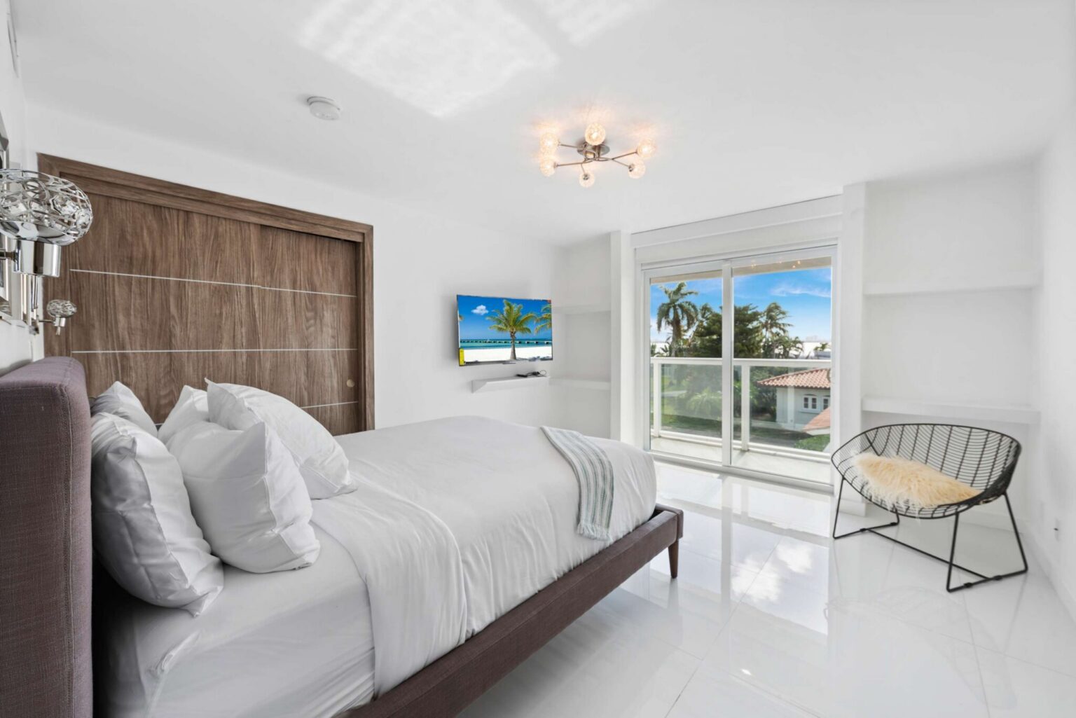 Bright guest bedroom featuring a queen-size bed with white linens, built-in closet storage, tile flooring, wall-mounted TV, contemporary lighting, and a large picture window providing natural light and views of the surrounding tropical neighborhood.