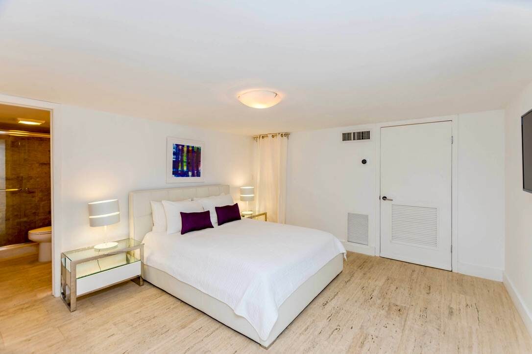 Guest bedroom suite featuring a queen-size bed with premium linens, bedside tables with lamps, wood-look flooring, overhead lighting, ensuite bathroom access, and a bright, functional layout designed for comfort and extended stays.