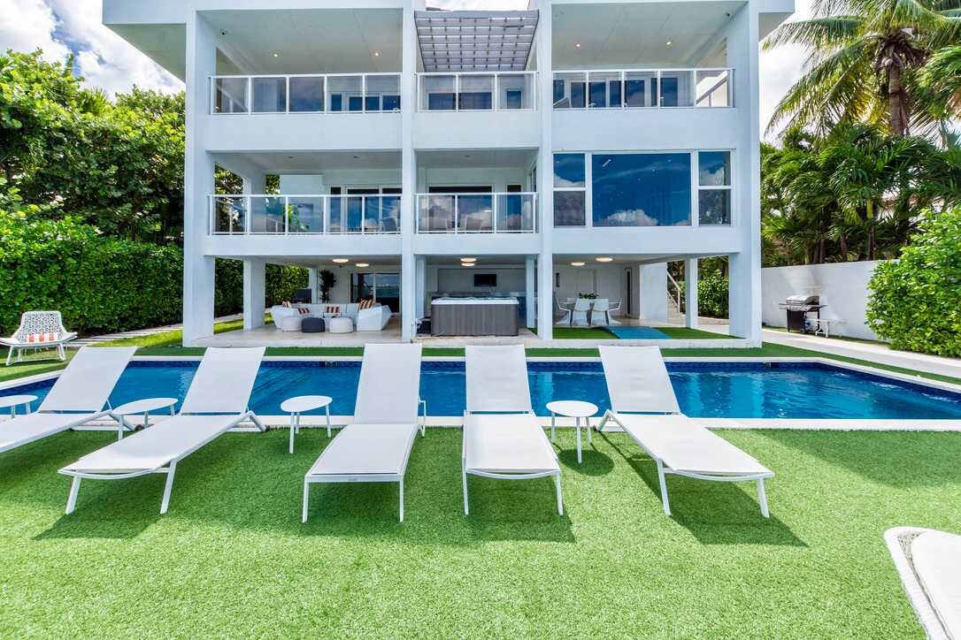 Three-story waterfront villa with a private swimming pool, sun loungers, landscaped lawn, covered outdoor living and dining areas, expansive balconies, floor-to-ceiling windows, tropical greenery, and direct waterfront views.