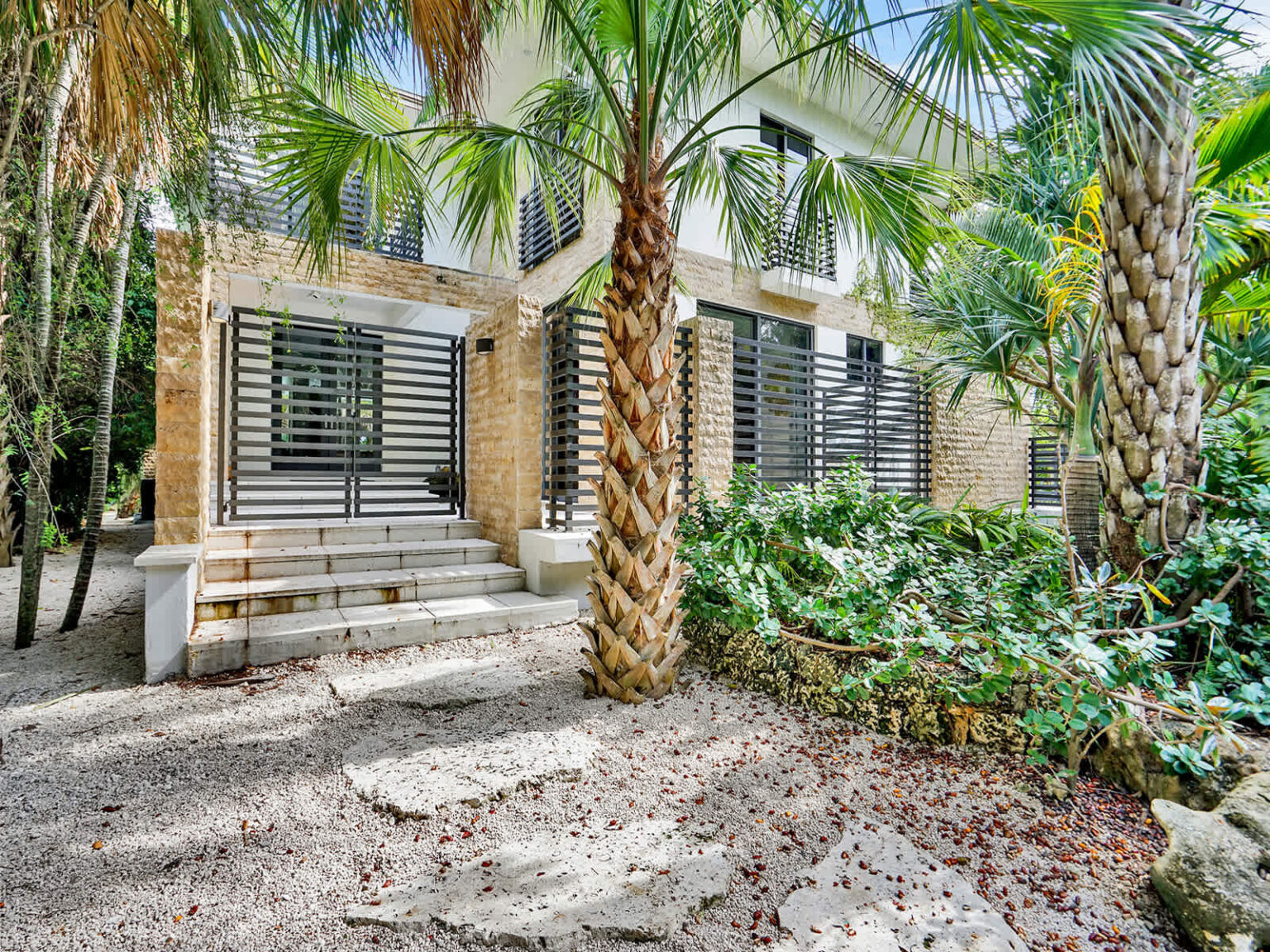 Tropical landscaped exterior featuring mature palm trees, native greenery, and a shaded ground-level seating area. Natural stone accents, modern architecture, and lush garden surroundings create a private and tranquil outdoor environment.