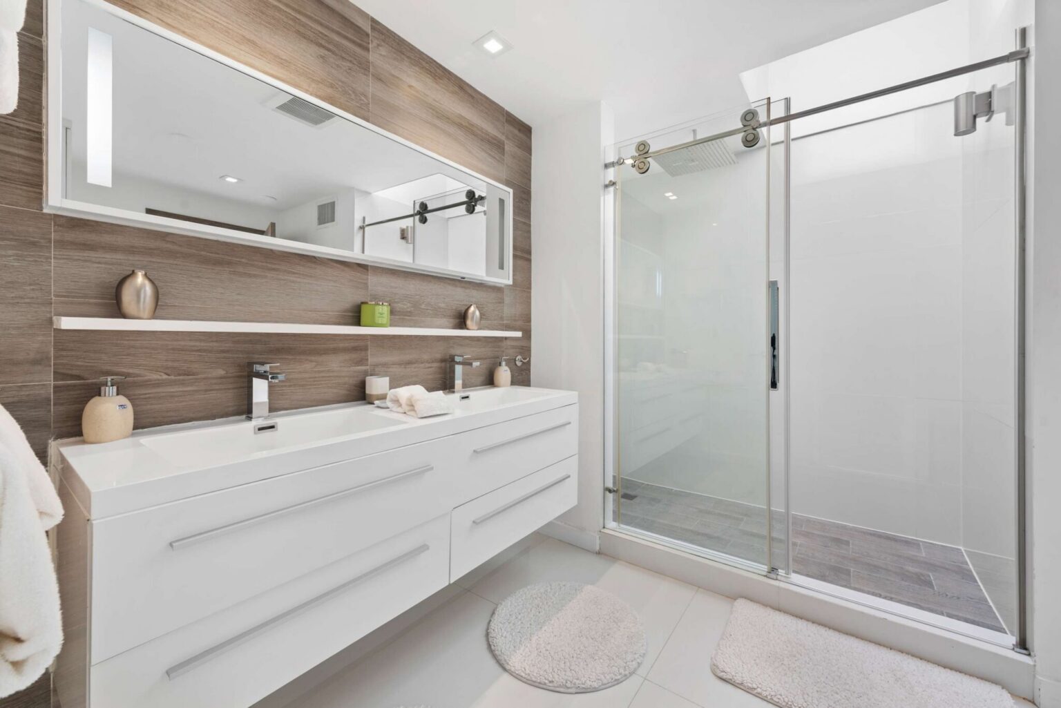Contemporary bathroom featuring a double-sink floating vanity, oversized wall mirror, glass-enclosed walk-in shower, wood-look accent wall, modern fixtures, built-in shelving, and a bright, functional layout with ample storage and counter space.
