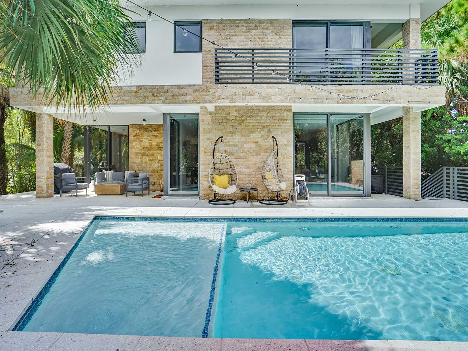 Contemporary outdoor space with a rectangular swimming pool, shallow lounge area, tiled deck, and direct villa access. Floor-to-ceiling glass doors connect indoor and outdoor living, surrounded by tropical landscaping.